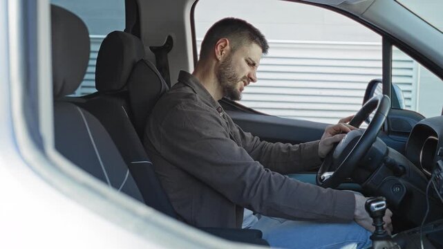 Man seated in car holding gearshift with one hand on the steering wheel while looking down beside a building; pensive thought.