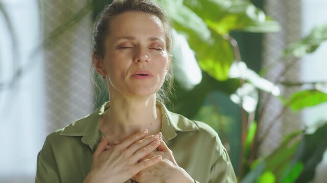 A woman takes a deep, calming breath with her hands clasped over her chest, surrounded by soft natural light and greenery, focusing on mental well-being and stress relief.