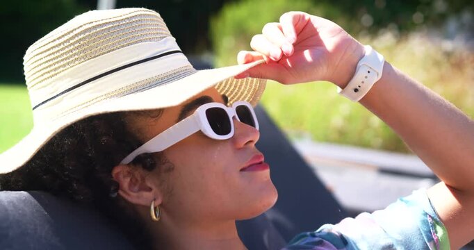 Adult female reclining on lounger raising straw hat brim shading eyes with white shades in sunlight