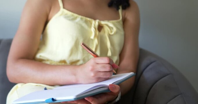 Female holding notebook on lap and lifting rose-gold pen, writing notes in home gray armchair