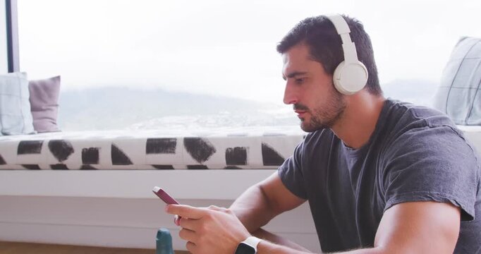 Adult man sitting right by window with headphones, glancing out, tapping phone, grabbing sunglasses