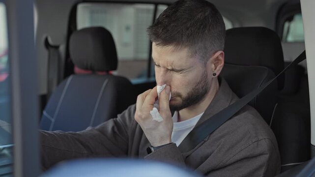 Man in car holds tissue to nose and winces while seated with seatbelt on, steering hand on wheel, parked on street; discomfort.