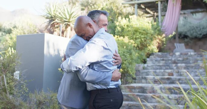 Diverse men greeting, man in blue shirt initiating hug, gray suit man sharing handshake on terrace