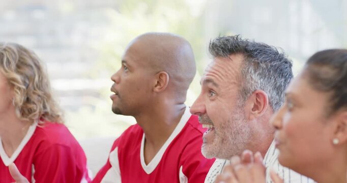 Diverse friends and family sitting on sofa watching off-screen play, two in red jerseys cheering