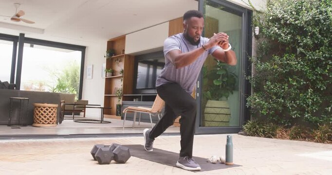 Shifting weight, African man doing curtsy lunges on patio mat beside weights, wrist tracker, toning