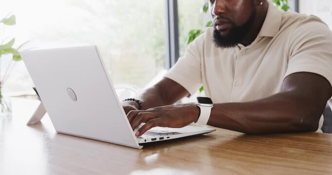Man typing on white laptop at wooden table by window with plants, wearing polo and smartwatch