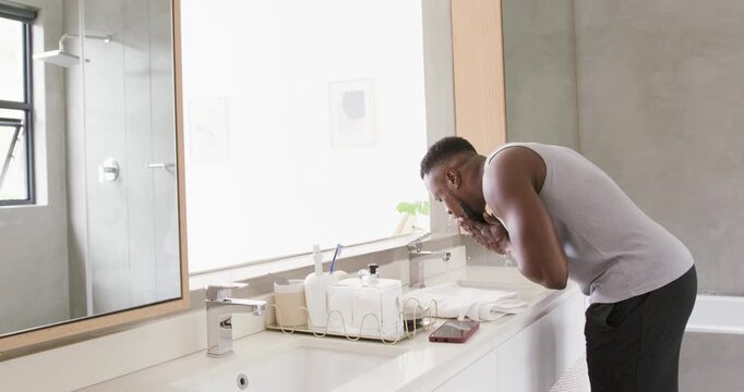African man turning on faucet at bathroom sink, washing face and beard, patting towel, copy space