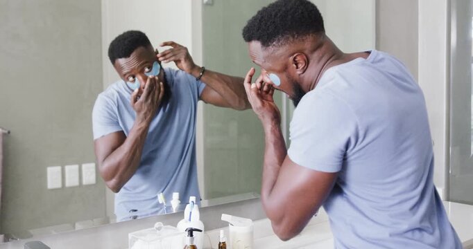 African American man in tee starting skincare, applying eye patches with mirror and reading phone