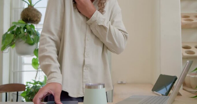 Non-binary worker taking call while typing on laptop, holding phone, jotting in notebook at home