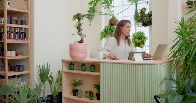 African nonbinary checking phone, typing on laptop, flipping planner, noting orders in plant shop