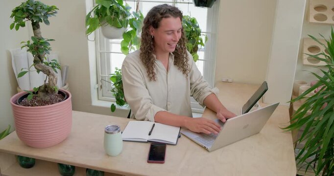 Nonbinary African American at desk seeing call, waving while typing on laptop and scrolling phone