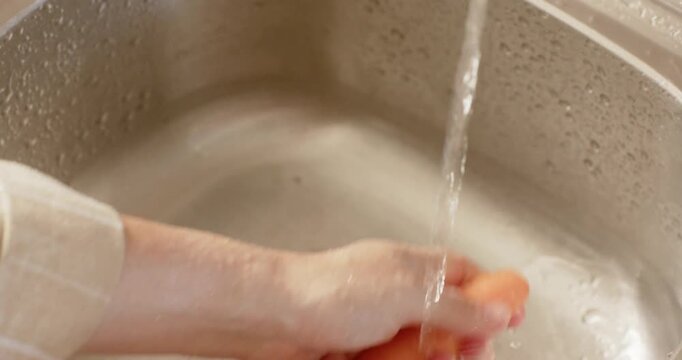Adult female hands wearing light sleeve rinsing and scrubbing carrot under running faucet at sink