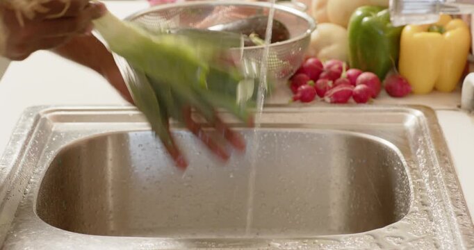 African American man holding leek at sink, rinsing, peeling leaves under running tap to remove dirt