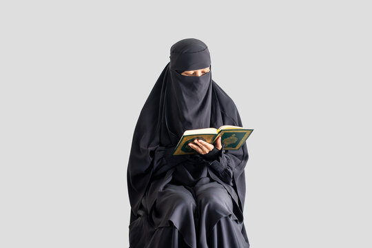 Woman in black niqab reads Quran calmly with plain light gray background. Focused expression shows devotion and peace. Ideal for themes of faith, study, or spiritual reflection.