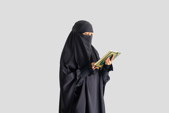 Woman in black niqab reads Quran calmly with plain light gray background. Focused expression shows devotion and peace. Ideal for themes of faith, study, or spiritual reflection.