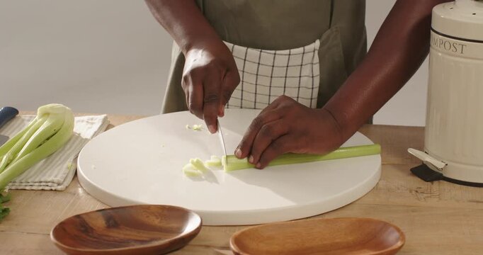 Mid-adult man slicing celery at kitchen counter on white board with chef's knife, wearing apron
