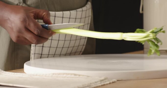 Man wearing checked apron chopping celery at kitchen counter on white cutting board with blue knife
