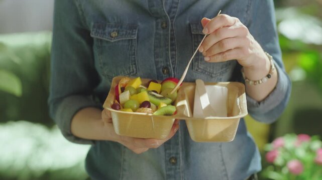 A woman uses a wooden fork to eat fresh fruit salad from a biodegradable container, emphasizing sustainable choices and healthy eating habits at home.