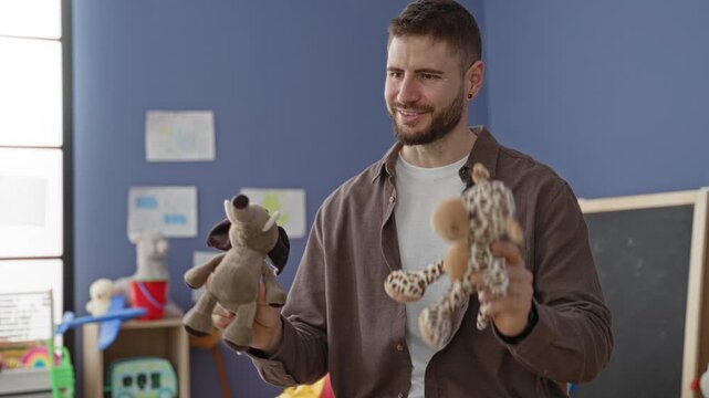 Young man holds two stuffed toys and puppets while smiling with open palms in a classroom building among shelves and a chalkboard; playful warmth.