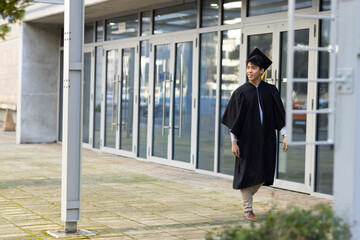 Asian man early 20s walking across plaza by glass doors wearing black gown mortarboard, copy space © wavebreak3