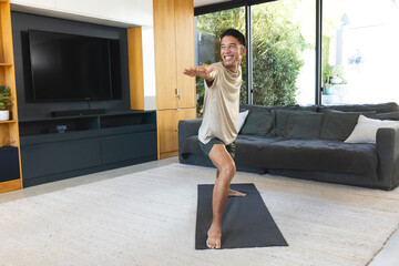 Adult man practicing standing yoga on mat at home near TV, wearing beige tee, dark shorts © wavebreak3