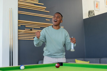 African American man shouting and clenching fist at green-felt table holding bottle, phone on rail © wavebreak3