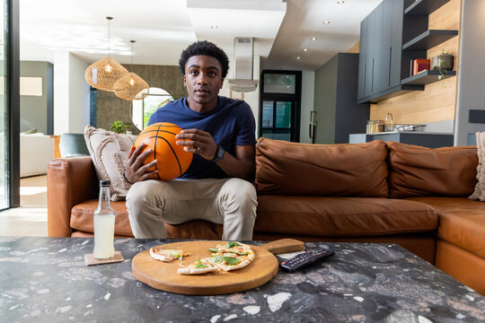 African American man sitting on sofa in living room holding basketball, wearing navy tee, khakis