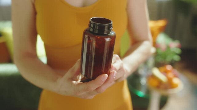 Close-up of a woman in yellow athletic wear pouring white vitamin capsules from a brown pill bottle into her hand as part of her daily wellness and nutrition routine.