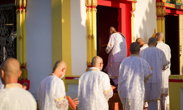 Thai men in white walk in line with palms together carrying folded orange monk robes as medium to large group approaches temple doorway, warm light before ordination.
