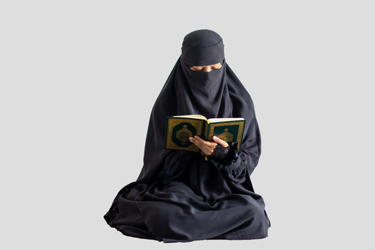 Woman in black niqab reads Quran calmly with plain light gray background. Focused expression shows devotion and peace. Ideal for themes of faith, study, or spiritual reflection.