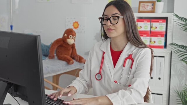 Hispanic woman doctor points finger while typing on computer with stethoscope in clinic building; professional care confidence.