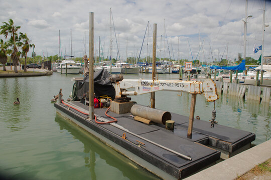 Rusty Crane Barge at Marina Dock With Boats and Palm Trees Under Cloudy Sky. Editorial Use Only, Gulfport, FL, USA, April 8, 2026. Sailboats in the background and pelicans on the water under a cloudy 