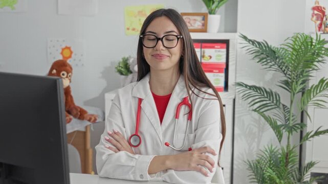 Hispanic woman doctor with stethoscope and glasses, arms crossed with hands visible at clinic building desk and computer; professional confidence.