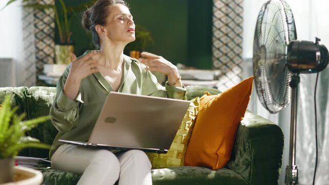 An adult woman sits on a green velvet couch with a laptop, visibly hot, fanning herself for relief from intense summer heat indoors. Captures the challenging reality of remote work during a heatwave.
