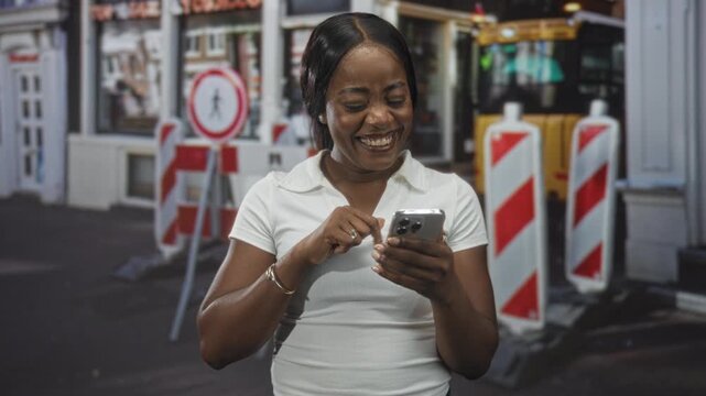 Woman smiling and tapping smartphone with fingers beside red white roadwork bollards and pedestrian sign on street; joy connection.