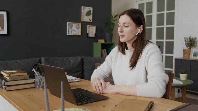 Medium shot of young Caucasian woman with earbuds sitting at desk while working from home, talking during online project meeting, suffering internet connection problems, checking and restarting router