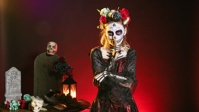 Creepy female model doing hush gesture to keep secret, wearing santa muerte costume and showing slience mute and secrecy sign with finger over lips. Holy dios de los muertos holiday.