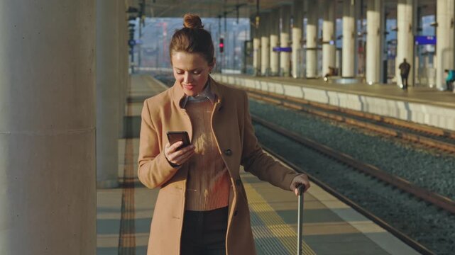 A happy woman in a light brown coat stands on a railway platform, smiling as she interacts with her smartphone while resting a hand on her suitcase handle.