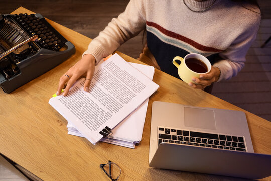Female screenwriter with coffee and script on table in office at night, top view