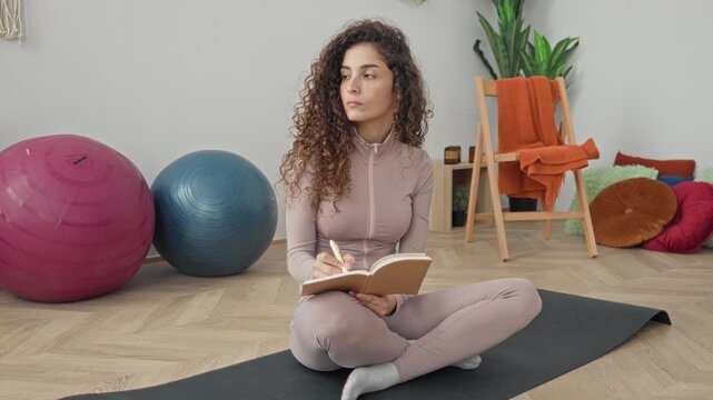 Woman writing in notebook on a yoga mat with two exercise balls and cushions visible in a bright home studio; mindfulness planning calm.