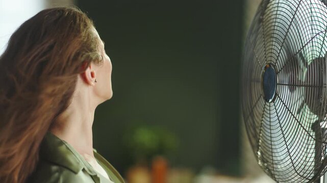 A content middle-aged woman with light brown hair finds refreshing relief as cool air from a large electric fan blows on her face and hair, smiling upwards in an indoor setting.