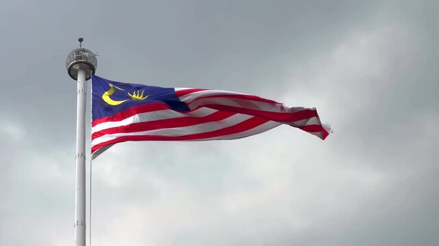 Flag of Malaysia waves proudly against cloudy sky. Tall pole holds flag high in wind. Colors vibrant, symbolizing national pride and unity. Suitable for patriotic, governmental, or cultural projects