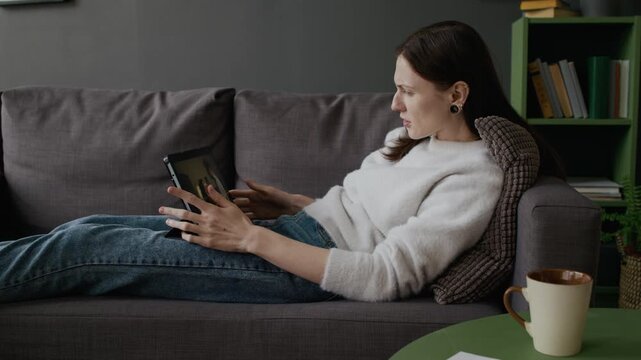 Medium full shot of young Caucasian woman resting on couch, watching movie on subscription streaming service app on digital tablet, frustrated and angry at internet connection interruption