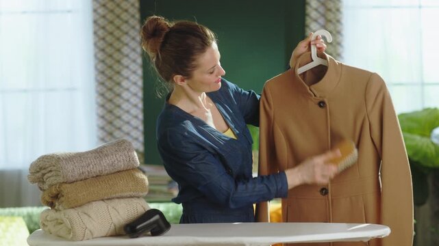 A smiling woman uses a clothes brush on a brown coat, with a fabric shaver and folded knitwear nearby, showcasing a thorough seasonal clothing maintenance routine.