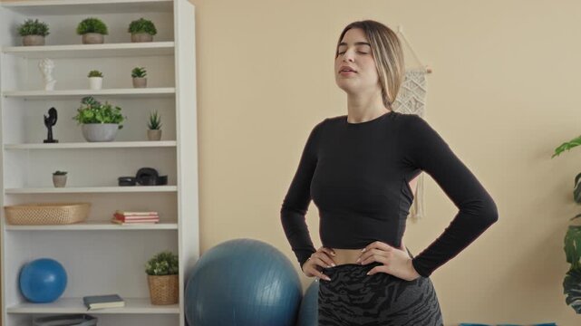Woman with hands on hips beside an exercise ball in a studio, standing with eyes closed taking steady breaths and holding a relaxed yoga pose; calm focus.