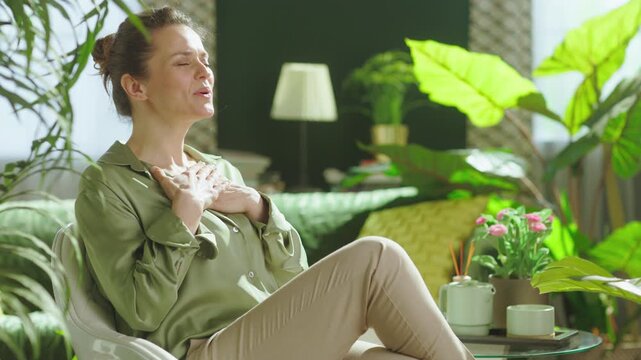A woman sits peacefully with her eyes closed and hands on her chest, practicing meditation and deep breathing in a warm, botanical home setting.