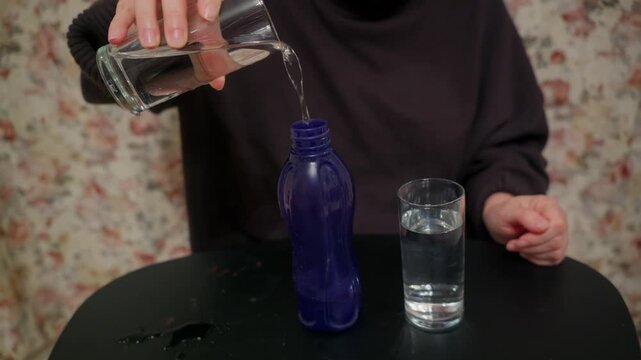 A person pours water from a glass into a blue reusable bottle, spilling some on the table. Another glass of water sits nearby. This highlights hydration, daily routines, and eco friendly habits