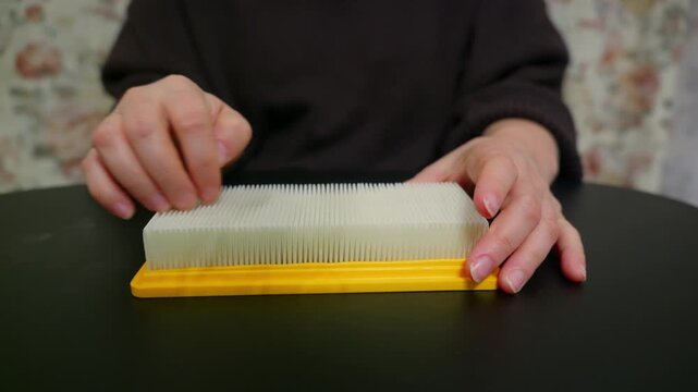 A persons hands gently stroke the pleated surface of a new rectangular car air filter with a yellow frame, creating a sensory experience. The close up shot focuses on the tactile interaction