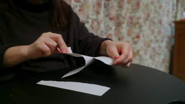 A close up of a woman's hands sitting at a black table. She repeatedly tears a long strip of white paper into smaller, uneven pieces, which fall onto the table surface