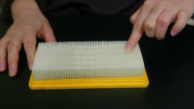 A woman's hands touch and inspect a new, clean rectangular engine air filter with a yellow frame, running her finger along the white pleats on a black table. Car maintenance concept